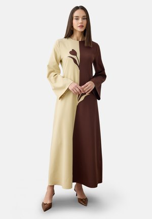 Mujer que lleva un vestido largo bicolor beige y marrón con diseño floral, mangas largas acampanadas y tacones marrones de punta estrecha.