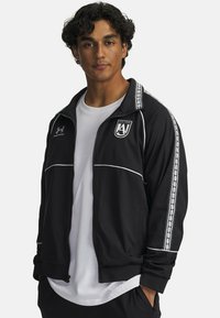 Black athletic jacket with white accents and side stripes. Features a zip front, stand collar, and embroidered logo on the chest.