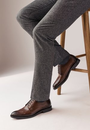 Chaussure habillée en cuir marron avec une finition lisse, design à lacets et bout rond. Pantalon gris texturé, assis de manière décontractée sur un tabouret en bois.