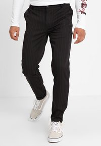 Man loopt en draagt een zwarte pantalon met krijtstrepen, een wit overhemd met lange mouwen en beige veter sneakers op een witte achtergrond.