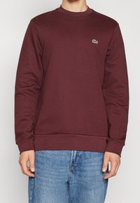 Sudadera burdeos de mezcla de algodón, con cuello redondo y puños acanalados. Pequeño logotipo verde en el lado izquierdo del pecho. Jeans visibles por debajo.