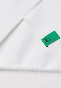 Tessuto bianco con sezioni a coste e strutturate, caratterizzato da un'etichetta verde con un'icona nera. Texture morbida evidente nel primo piano.