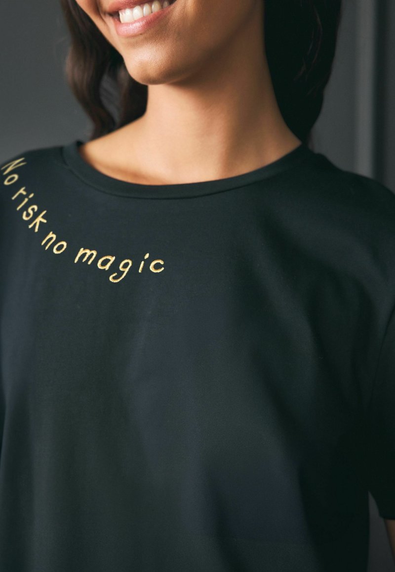 Donna sorridente che indossa una maglietta nera con la scritta dorata "No risk no magic" curva sull'area della spalla.