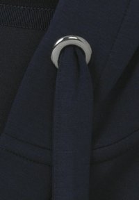 Donkerblauwe stof met een gladde textuur, voorzien van een metalen cirkelring die een stoffen band vasthoudt. Het ontwerp benadrukt eenvoud en functionaliteit.