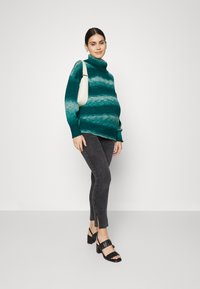 Pleteni teal pulover s vodoravnimi črtami, s polo ovratnikom in rebrastimi manšetami, v kombinaciji s temno sivo oprijetimi hlačami in črnimi sandali na peto.