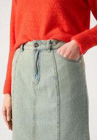 Jeanskje i lyseblått med knappestramming foran, har sidelommer og vertikale sømdetaljer, parret med en rød strikkegenser.