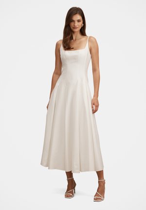 Žena v bielych midi šatách bez rukávov so štvorcovým výstrihom, priliehavým živôtikom a rozšírenou sukňou, doplnených bielymi sandálmi na podpätku.