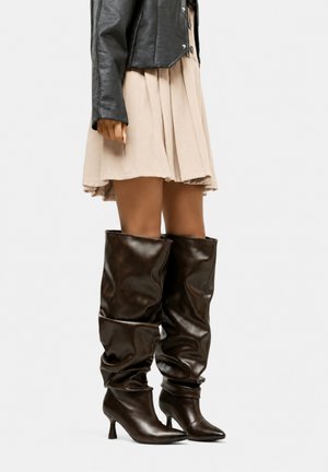 Femme portant des bottes en cuir marron foncé, décontractées, montantes jusqu'au genou, à bouts pointus et talons fins, associées à une jupe beige et une veste noire.