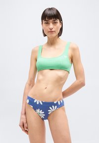 Femme portant un crop top en maille verte et un bas de bikini bleu avec des imprimés marguerites blanches, debout avec une main derrière le dos.