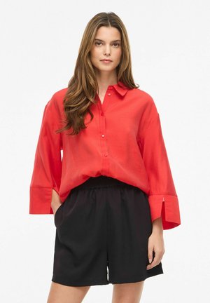 Femme aux longs cheveux bruns portant une chemise rouge ample boutonnée et un short noir taille haute, debout avec une main dans la poche.