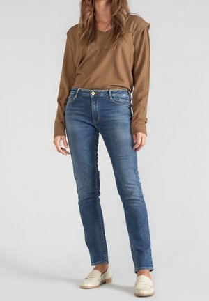 Henkilö, jolla on päällään ruskea pitkähiha-paita, siniset slim-fit-farkut ja luonnonvalkoiset loafersit seisoo yksivärisen taustan edessä.