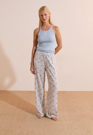 Femme blonde debout pieds nus portant un haut sans manches bleu clair et un pantalon blanc ample avec de petits motifs croissants orange.