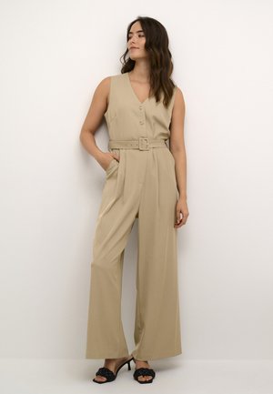 Beige jumpsuit med V-udskæring, knapfront og brede ben. Inkluderer et matchende bælte i taljen. Modellen har sorte hæle sandaler på.