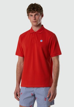 Uomo che indossa una polo rossa a maniche corte con un piccolo logo circolare blu e bianco, e pantaloncini a righe blu e bianche con motivi di corallo rosso.