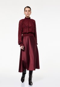 Top de encaje burdeos con escote y puños acampanados, combinado con una falda de satén burdeos y botas negras brillantes hasta la rodilla.