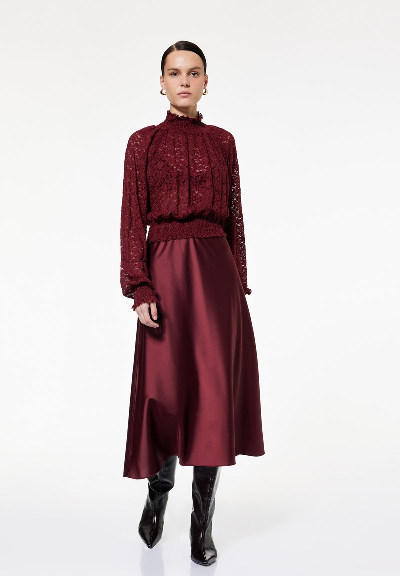 Top de encaje burdeos con escote y puños acampanados, combinado con una falda de satén burdeos y botas negras brillantes hasta la rodilla.