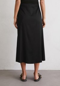 Falda larga negra en línea A, hecha de un tejido suave, con un brillo sutil y una silueta fluida, combinada con sandalias planas negras.