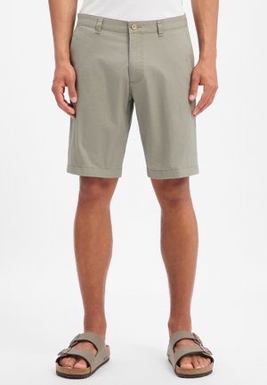 Mann trägt hellgraue knielange Shorts, weißes Hemd und graue Sandalen mit Doppelriemen und steht vor einem weißen Hintergrund.
