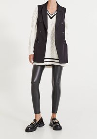 Czarna kamizelka dwurzędowa na białym swetrze w warkocze z czarnymi paskami, w zestawie z błyszczącymi czarnymi legginsami skórzanymi i mokasynami.