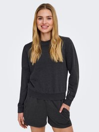 Jeune femme aux longs cheveux blonds portant un sweat-shirt gris foncé et un short assorti, souriant avec une main dans la poche.