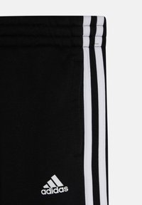 Zwarte trainingsbroek met drie witte strepen aan de zijkanten, met een klein wit Adidas-logo aan de voorkant. Zacht, sportief textuur van de stof.