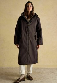 Sieviete, kas ģērbta tumši brūnā garā mētelī ar kapuci, baltās biksēs, bežās zeķēs un brūnās mokasīnās, stāv uz vienkāršas bēšas sienas fona.