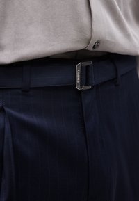 Παντελόνι μπλε ναυτικού με λεπτές ρίγες και μαύρη ζώνη με ασημένια ορθογώνια αγκράφα χαραγμένη με "Calvin Klein." Μπεζ πουκάμισο μέσα.