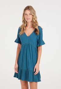 Teal mekko, jossa on V-pääntie, lyhyet röyhelöillyt hihat ja kerroksellinen helma. Rypytetty yksityiskohta rinnan alla ja kevyt materiaali.