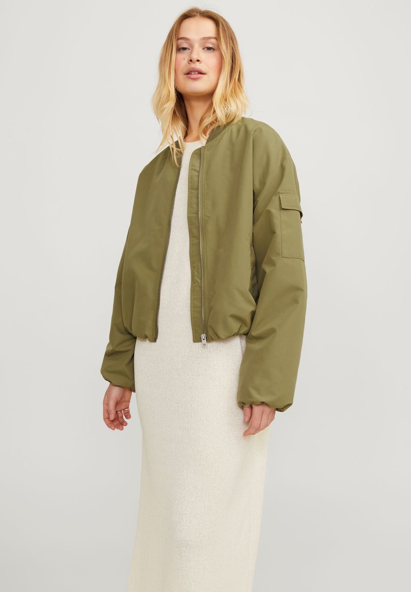 Jaqueta bomber verde-oliva com fecho de correr, punhos canelados e um bolso lateral, combinada com um vestido maxi em creme texturizado.