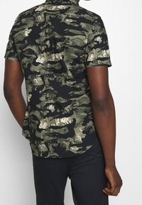 Korte mouwen blouse met camouflagepatroon, bestaande uit donker groen, zwart en beige kleuren met een gestructureerde stof en een puntige kraag.