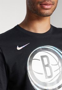 T-shirt en coton noir avec un grand graphique en argent représentant un ballon de basket et un logo Nike holographique sur l'épaule gauche. Encolure ronde.