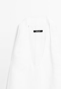 Haut en lin blanc avec un décolleté en V profond, portant une étiquette "Massimo Dutti". Le tissu a une texture lisse avec peu de plis.