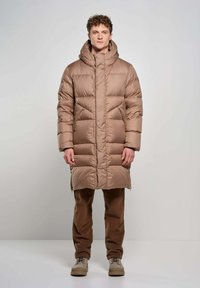 Gevoerde bruine lange jas met een hoge kraag en een oversized capuchon, voorzien van horizontaal gekwilt delen en zijzakken. Ggedragen met bruine corduroy broek.