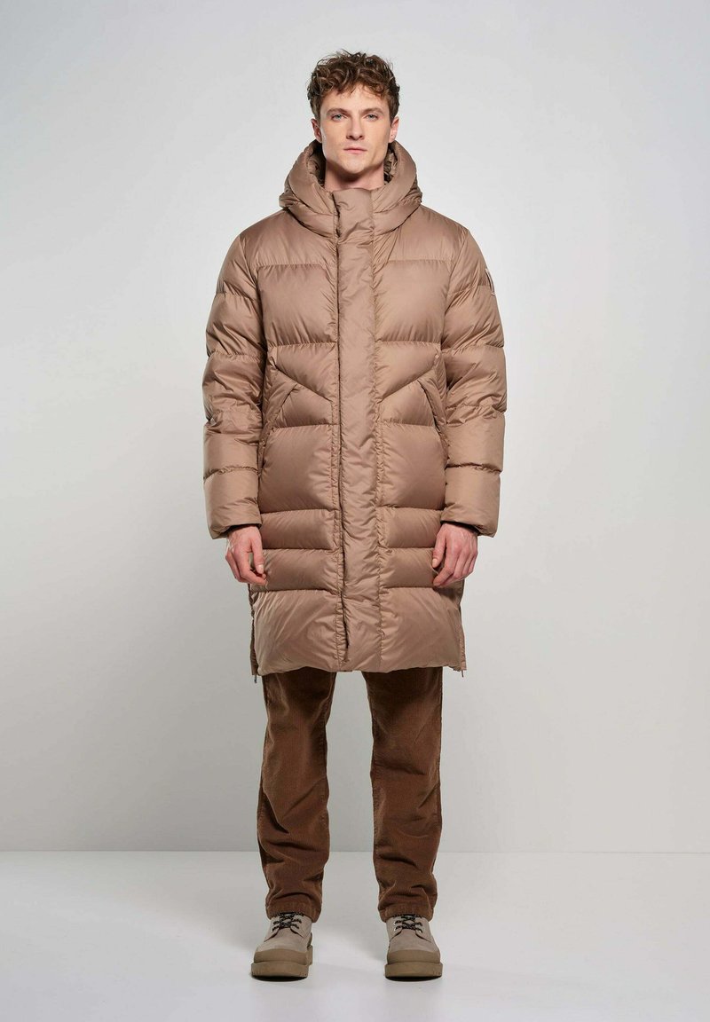 Gevoerde bruine lange jas met een hoge kraag en een oversized capuchon, voorzien van horizontaal gekwilt delen en zijzakken. Ggedragen met bruine corduroy broek.
