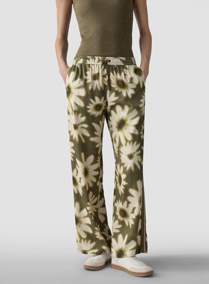 Modell trägt olivgrüne, weit geschnittene Hosen mit großem weißem und gelbem Blumenmuster, kombiniert mit olivgrünem Tanktop und weißen Sneakers.