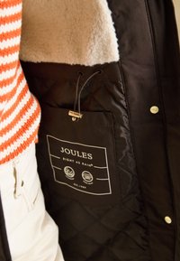 Veste noire avec intérieur matelassé, dotée d'un col en polaire beige et d'une étiquette avec le logo "JOULES". Cordon de serrage réglable à l'intérieur.