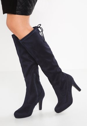 Kobieta nosząca granatowe zamszowe botki na obcasie sięgające do kolan z bocznym zamkiem błyskawicznym i sznurowaniem z tyłu, zestawione z czarną pikowaną spódnicą.