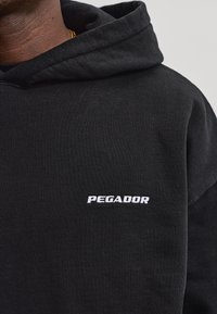 Nahaufnahme einer Person, die einen schwarzen Kapuzenpullover mit dem weißen Schriftzug "PEGADOR" auf der Brust trägt.