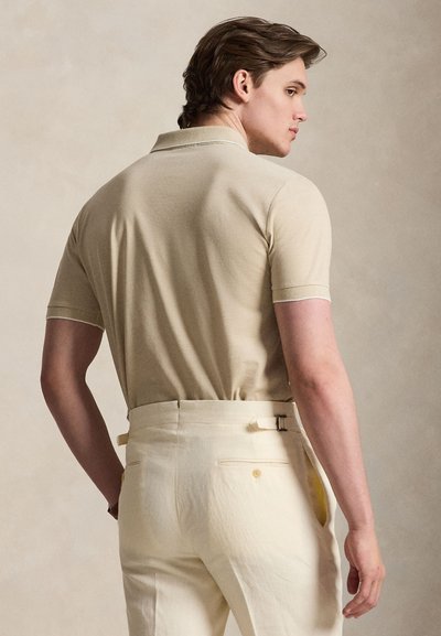 Άνδρας που φοράει μπεζ πόλο με κοντά μανίκια, μπαίνει σε κρεμ παντελόνια με ψηλή μέση και ρυθμιζόμενους ιμάντες στα πλάγια, στέκεται μπροστά σε απλό φόντο.