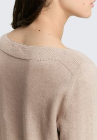 Vrouw in een beige gebreide trui, gedeeltelijk gekeerd om de nek en schouder te onthullen met twee kleine moedervlekken zichtbaar op de huid.