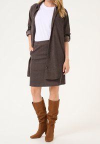 Kvinde iført en hvid t-shirt, en mørkebrun silkeskjorte med opkrøllede ærmer, matchende nederdel og løse brune ruskindsstøvler.