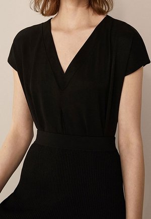 Femme portant un haut noir à manches courtes et col en V rentré dans une jupe noire côtelée taille haute sur un fond neutre.