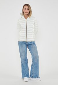 Giacca bianca imbottita con zip frontale e cappuccio attaccato, abbinata a jeans svasati blu chiaro e sneakers bianche. Texture liscia, design minimale.