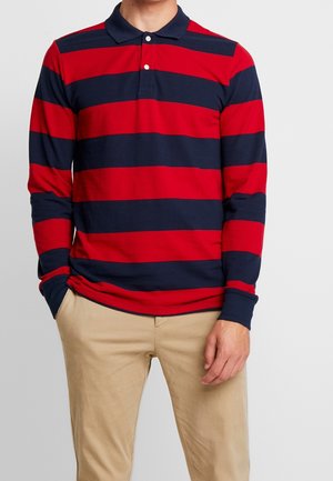 Mann trägt ein rot und marineblau horizontal gestreiftes Langarm-Poloshirt mit geknöpftem Kragen und beige Hose, Hände entspannt seitlich.