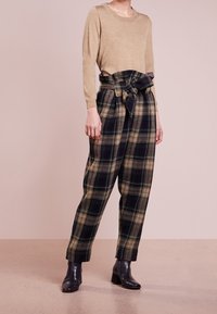 Femme portant un haut beige à manches longues, un pantalon taille haute à carreaux avec ceinture nouée à la taille, et des bottines noires, debout devant un fond uni.