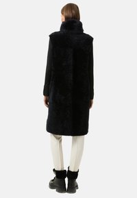 Granatowy kamizelka z futra syntetycznego z wysokim kołnierzem, detalami szwu na plecach i gładką fakturą, połączona z kremowymi wąskimi spodniami oraz czarnymi botkami za kostkę z futrzanymi akcentami.