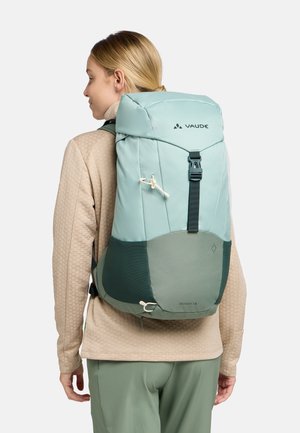 Zaino Vaude azzurro chiaro presenta un coperchio superiore, tessuto testurizzato, design bicolore e spallacci regolabili. Il modello indossa una maglia beige a fantasia.