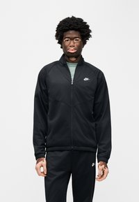 Μαύρο μπλουζάκι Nike με φερμουάρ και ψηλό γιακά, με λευκό λογότυπο στο αριστερό στήθος. Συνδυάζεται με ταιριαστό μαύρο παντελόνι, με λεία υφή.