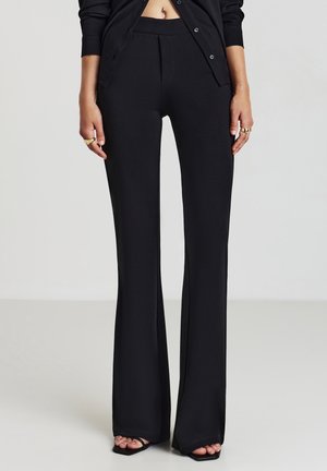 Vrouw die zwarte hoge taille, uitlopende broek draagt en een zwarte, met knopen gesloten cardigan, met ringen en zwarte sandaal met open tenen, staand op een lichte vloer.