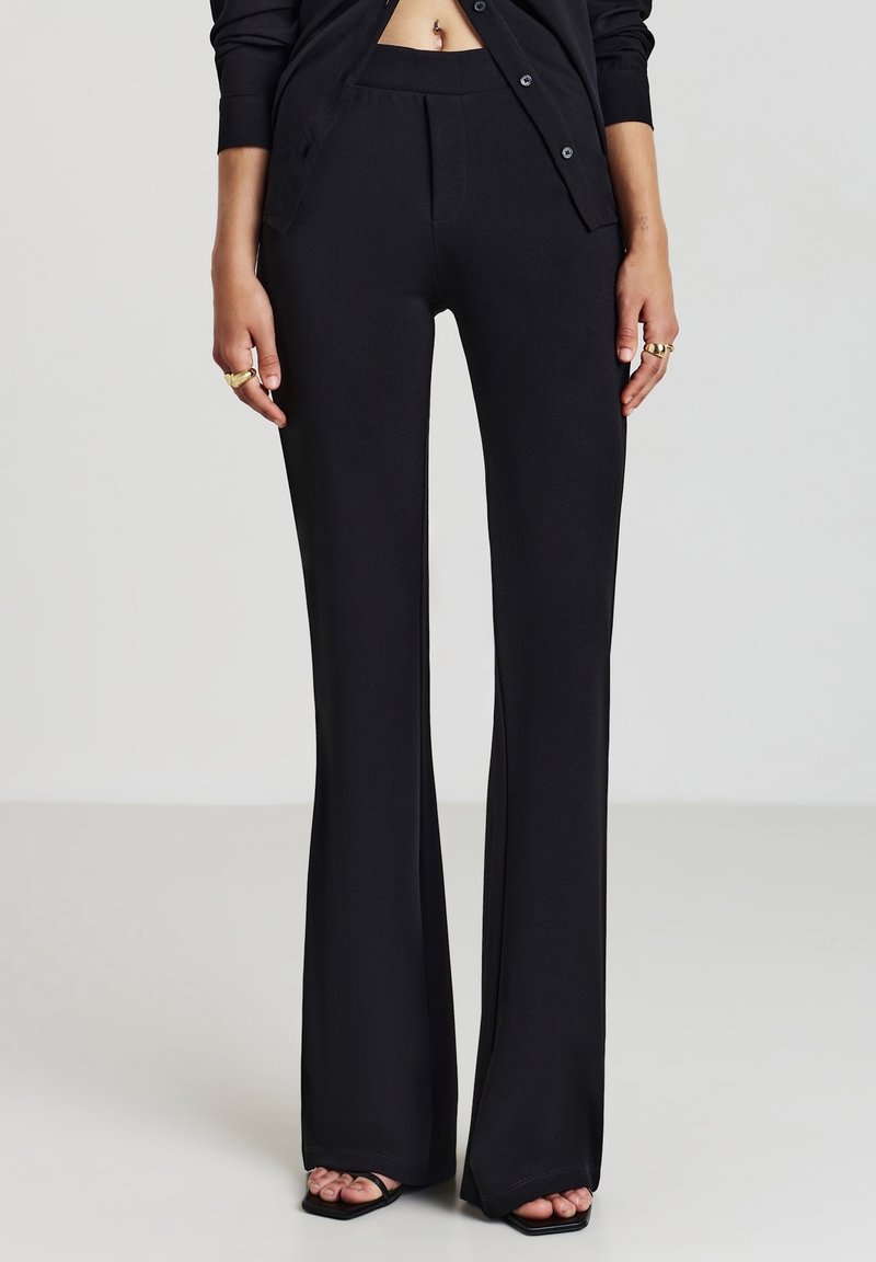 Femme portant un pantalon noir taille haute évasé et un cardigan noir boutonné, avec des bagues et des sandales noires ouvertes aux orteils, debout sur un sol clair.